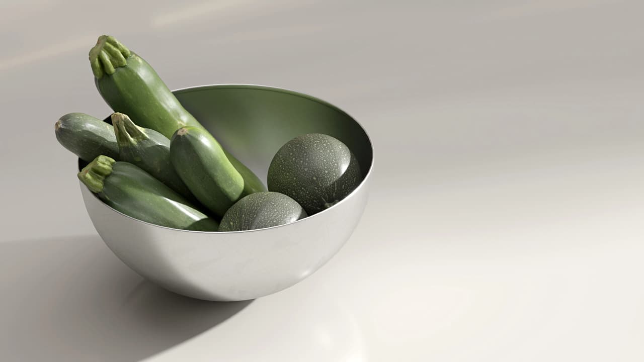 A minimalist close-up of a matte silver 3d bowl filled with vibrant green zucchini and round squash. The bowl sits on a smooth, light grey surface, casting a soft shadow under bright studio lighting.