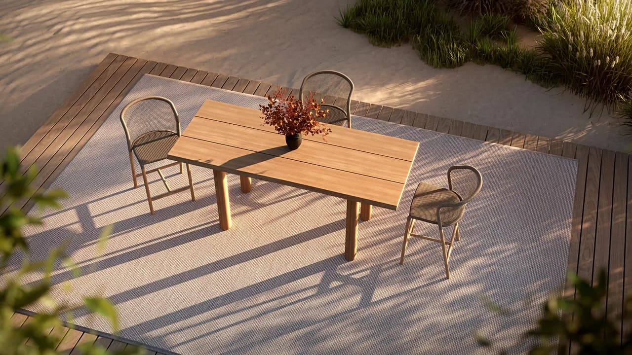 A top-down view of an cgi outdoor dining set featuring a wooden table and three woven chairs placed on a light textured rug over a wooden deck. The scene is framed by sand dunes and wild coastal grass, with warm golden hour sunlight casting long, dramatic shadows across the patio.