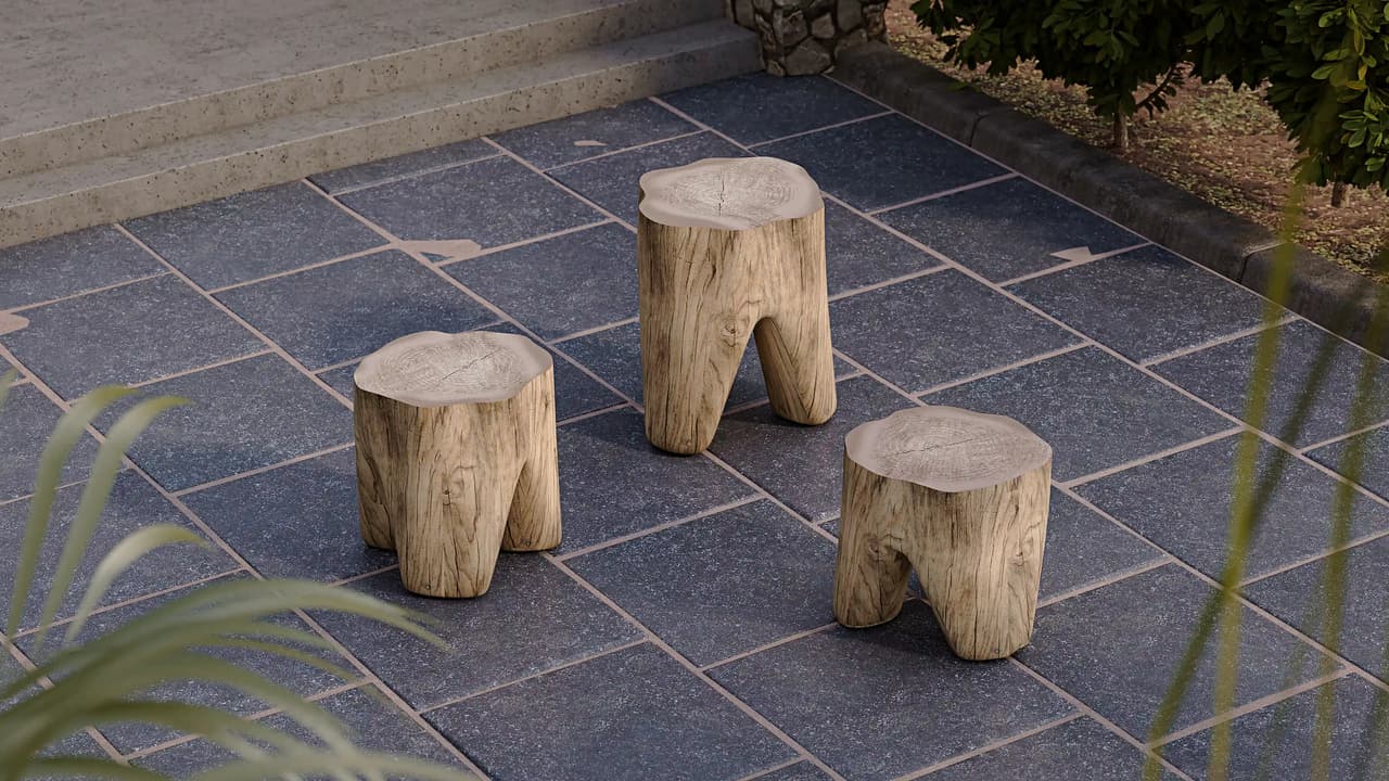 An outdoor patio scene featuring three rustic cgi stools carved from light wood logs with thick, root-like legs. They rest on dark square stone pavers, framed by softly blurred green foliage in the foreground under bright natural daylight.