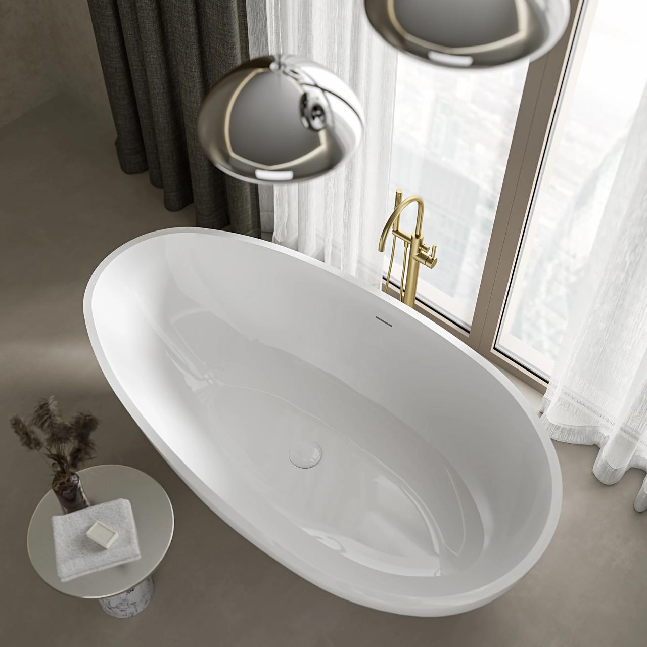 A high-angle view of a smooth white oval freestanding 3d bathtub placed on a grey tiled floor near a large window with sheer white curtains. The setup includes a floor-mounted brushed gold faucet, chrome pendant lights above, and a small side table with folded towels and soap.