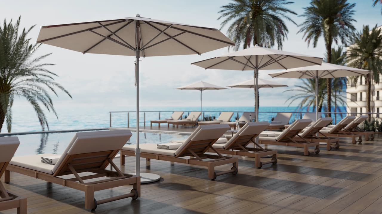 Low-angle perspective of white patio umbrellas and wooden sun loungers arranged on a timber deck by an infinity pool overlooking the ocean and palm trees