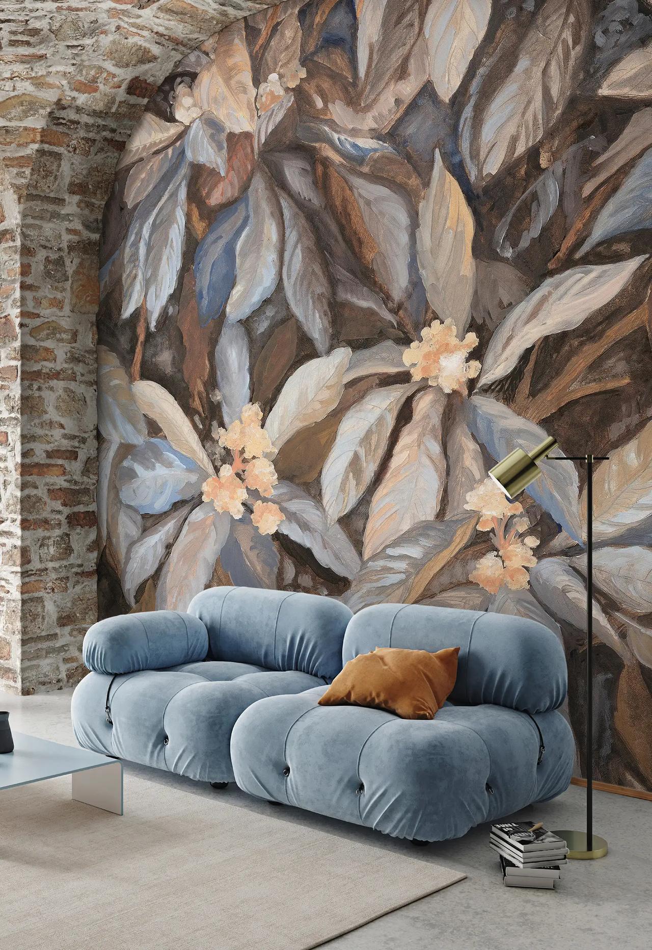 A dramatic 3d wall mural featuring painted oversized leaves and delicate peach flowers covers the main wall next to an exposed brick arch. A plush light blue modular sofa sits in the foreground, perfectly framing the earthy tones of the wallpaper.