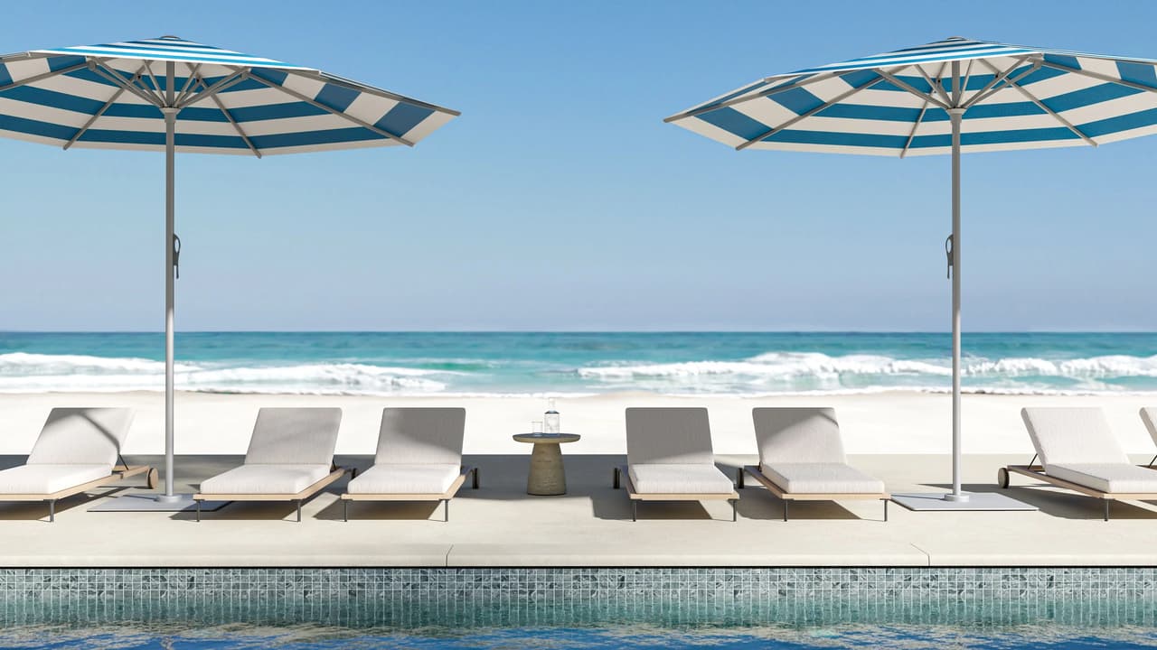 Frontal view of blue and white striped patio umbrellas with modern sun loungers arranged along a luxury resort pool overlooking a white sand ocean beach