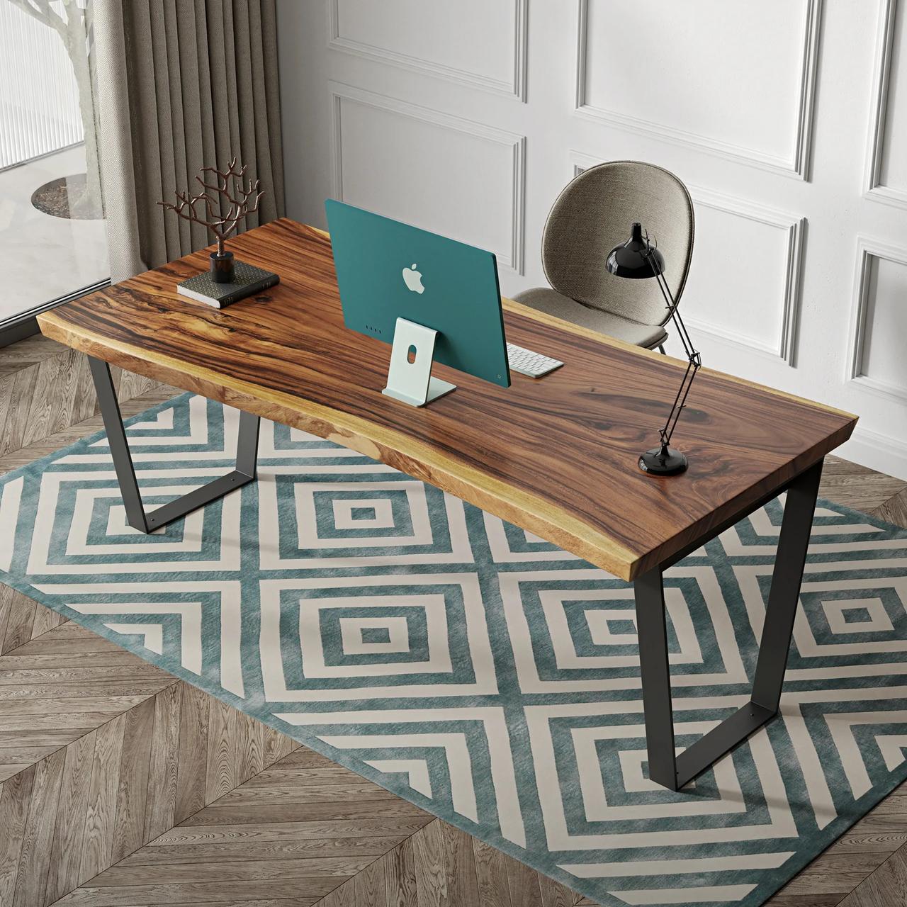 A top-down perspective of a stylish home office. A live-edge wooden desk with black metal U-shaped legs sits on a teal and white geometric rug. On the desk is a green computer monitor, a black lamp, and a small decorative tree, with a grey chair and white paneled walls in the background.