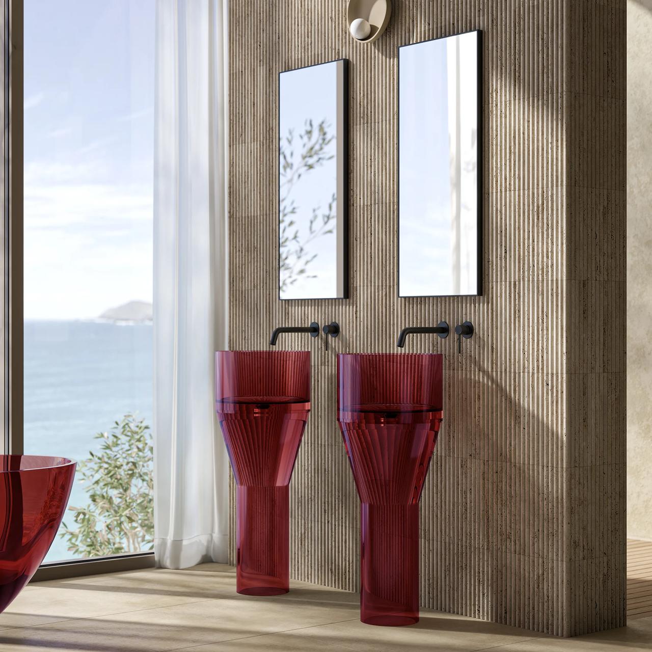 A luxurious, sunlit 3d bathroom featuring twin translucent ruby-red pedestal sinks with a ribbed texture, set against a matching fluted wooden wall. The setup includes two tall rectangular mirrors, wall-mounted black faucets, and a large floor-to-ceiling window offering a serene ocean view.