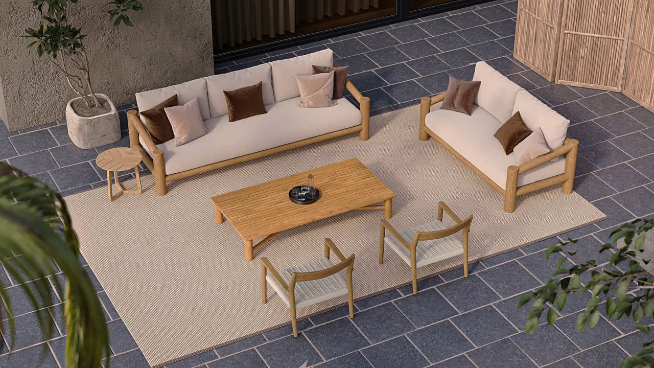 An isometric high-angle view of a luxurious 3d outdoor lounge area on a paved terrace. A complete wooden-framed furniture set, including a 3-seater sofa, a 2-seater sofa, and two woven cane armchairs with beige cushions, is arranged around a central slatted coffee table on a large woven area rug. Assorted decorative pillows in chocolate brown, terracotta, and patterned grey add warmth. A potted tree in a concrete planter and a blurred palm frond frame the scene against a modern house wall with vertical wood slats. The scene is bathed in warm, natural daylight casting distinct shadows.