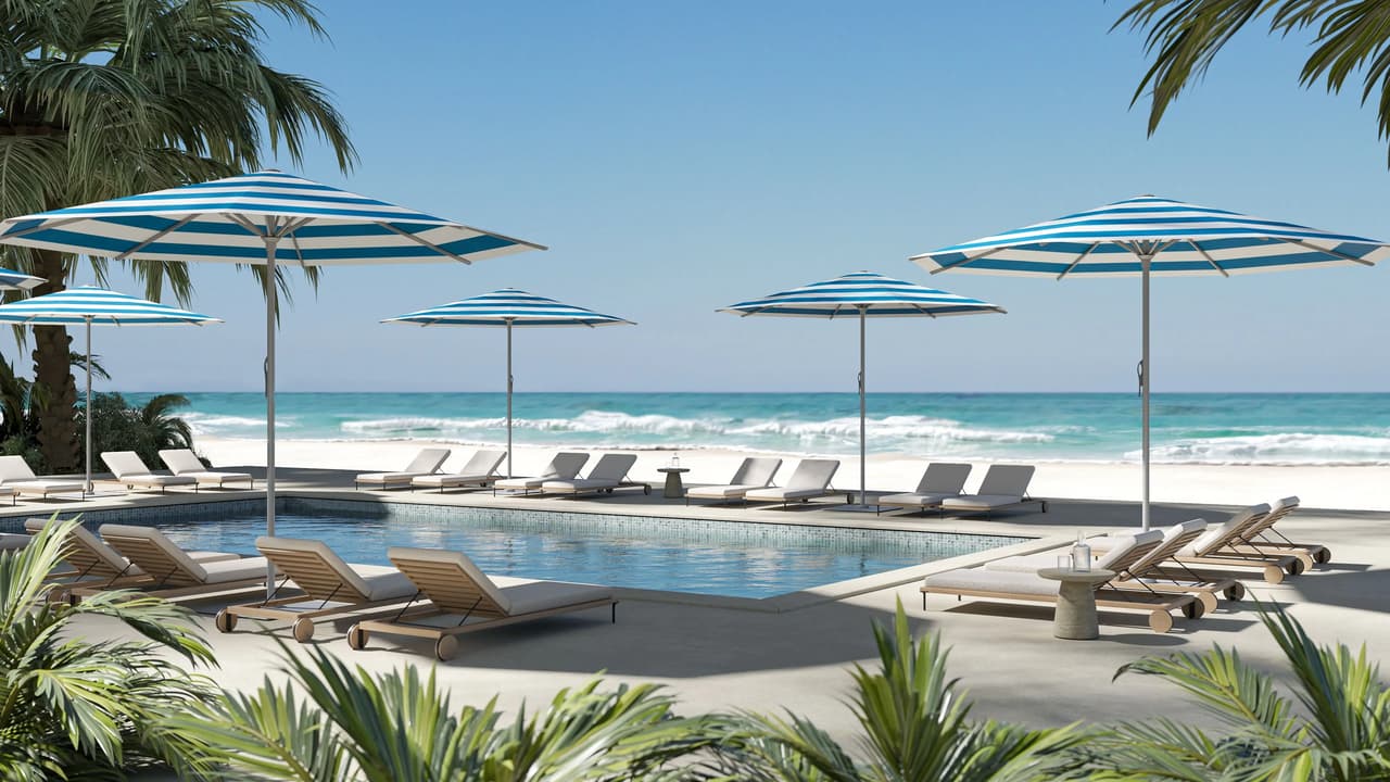Wide view of a luxury resort pool deck lined with blue and white striped patio umbrellas, wooden lounge chairs, and tropical palm trees overlooking a ocean beach