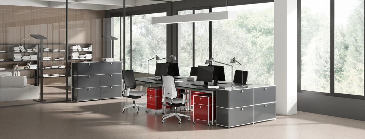 A spacious, sunlit open-plan cgi office with large windows overlooking green trees. The central workspace features a long shared desk, ergonomic chairs, and striking red and grey modular metal cabinets, separated from a cozy lounge area by a sleek glass partition.