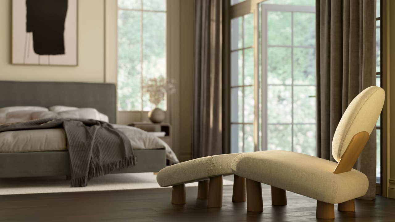 A shallow depth of field shot focusing on a unique cream lounge 3d chair with thick wooden legs and a matching footstool. The softly blurred background reveals a modern bedroom with a grey upholstered bed, a draped blanket, and a large window with brown curtains.