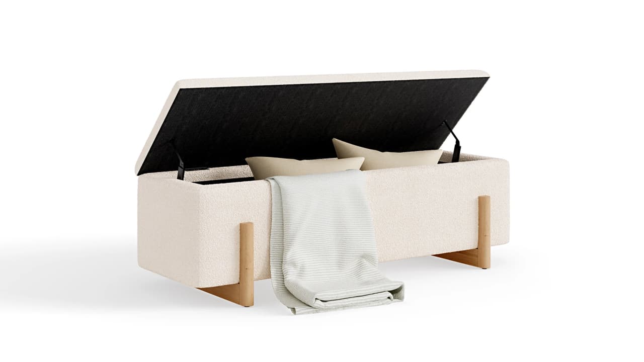 An isolated 3D product render of an upholstered cream boucle storage ottoman with a raised top showing stored pillows inside. The bench rests on natural wooden legs with a soft textured throw blanket draped over the front edge against a solid white background. 