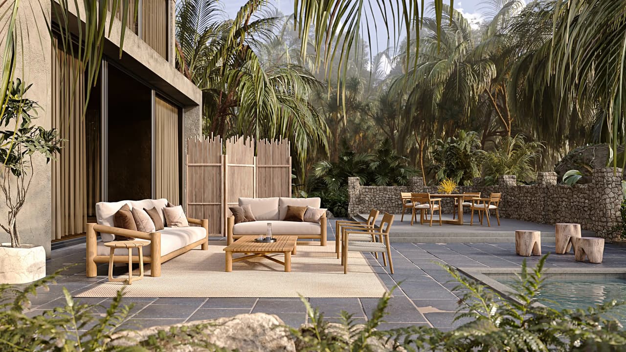 A spacious outdoor terrace paved with dark stone features a modern wooden lounge set with cream cushions on a light rug. The scene is surrounded by lush tropical palm trees, a rustic stone wall, a dining area in the background, and a swimming pool on the right under bright sunlight.