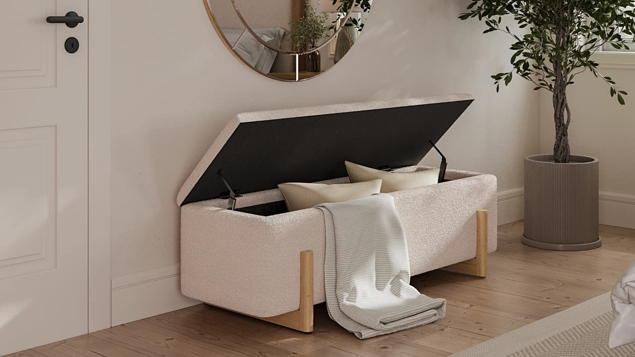 A white cgi textured storage bench with light wooden legs sits on a wooden floor, its lid open to reveal two pillows inside while a striped blanket drapes over the edge. The cozy room features a white paneled door, a round wall mirror, and a tall potted plant.