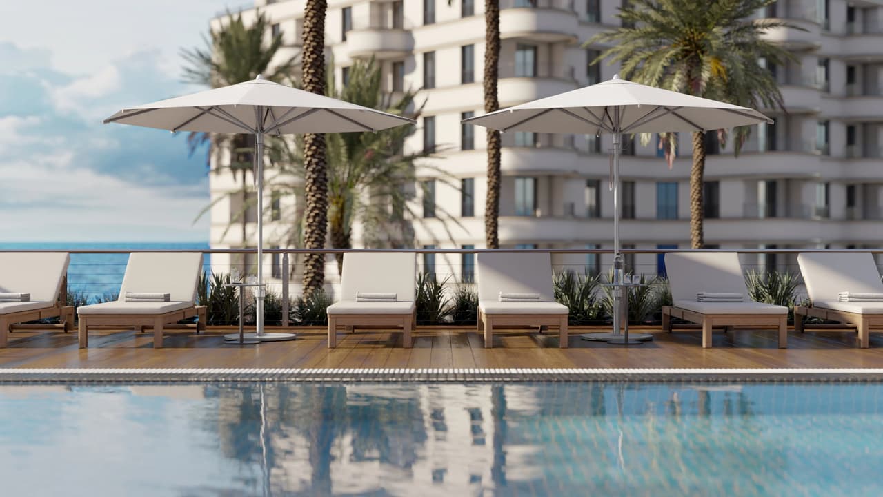 A sunny outdoor resort scene with a swimming pool in the foreground. A wooden deck is lined with wooden sun loungers featuring thick white cushions and small side tables, shaded by two white patio umbrellas. Palm trees, a glass railing overlooking the ocean, and a large white multi-story hotel building complete the background.