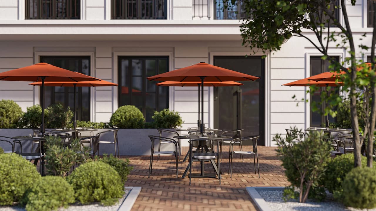 Frontal view of an outdoor restaurant terrace with terracotta orange patio umbrellas, round dining tables, black metal chairs, and decorative green bushes along a paved courtyard