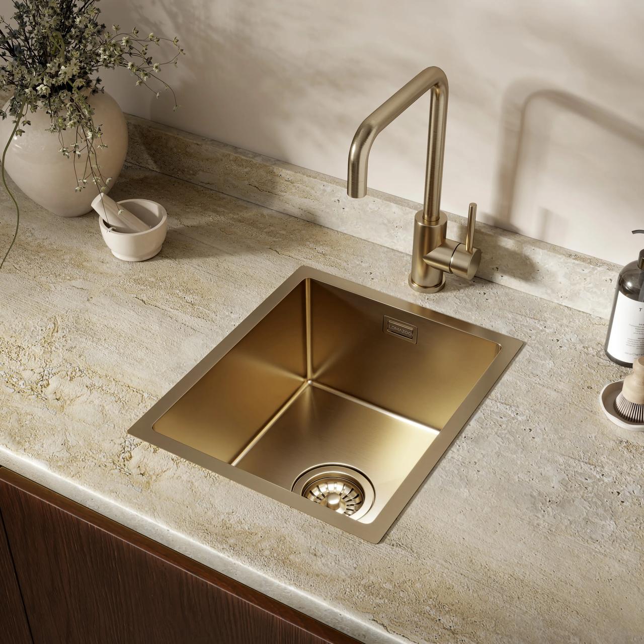 Close-up of a brushed gold square kitchen sink and matching brass faucet integrated into a light textured stone countertop