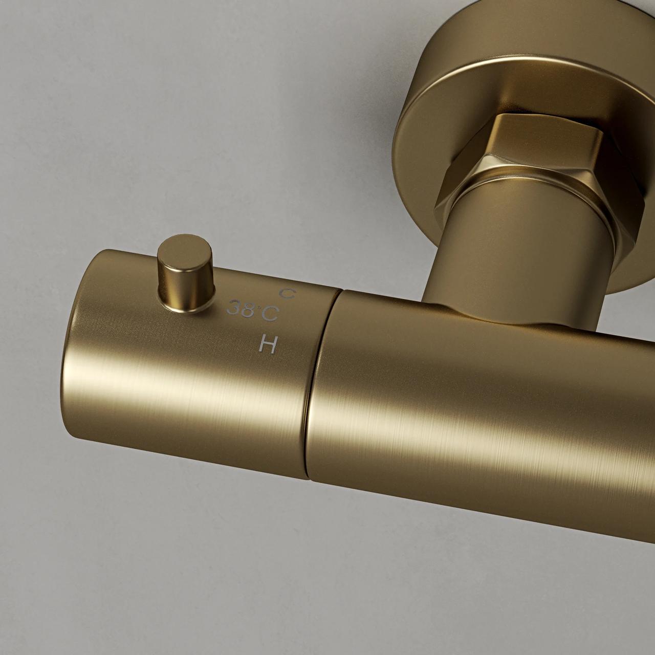 A detailed macro shot of a wall-mounted brushed gold 3d shower mixer against a light grey background. The close-up highlights the matte metallic texture, a temperature safety button, and subtle engraved temperature markings.