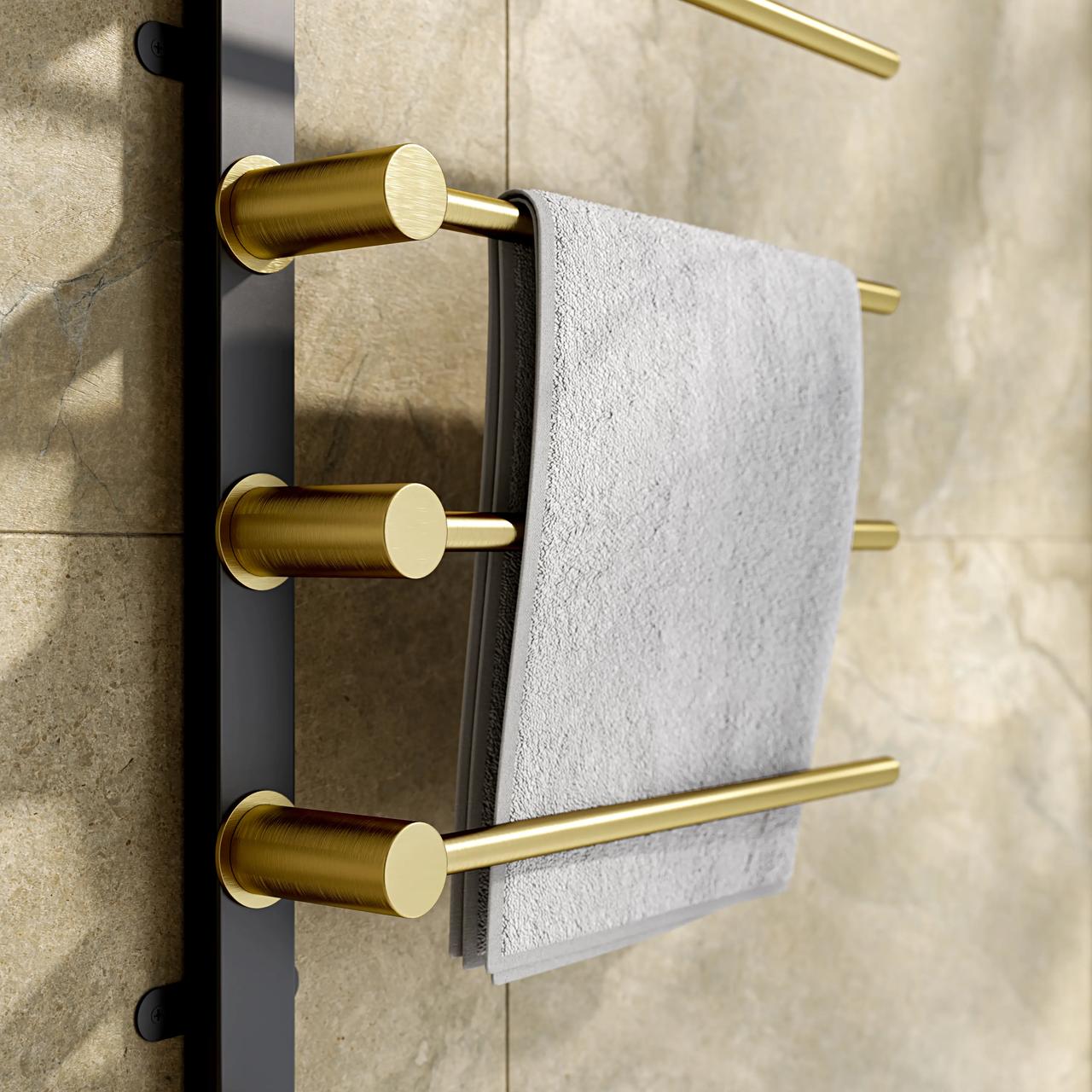 A close-up shot of a sleek electric 3d towel warmer with cylindrical brushed gold bars attached to a vertical matte black frame. A soft, textured white towel is draped over one of the heated bars against a warm beige stone tile background.