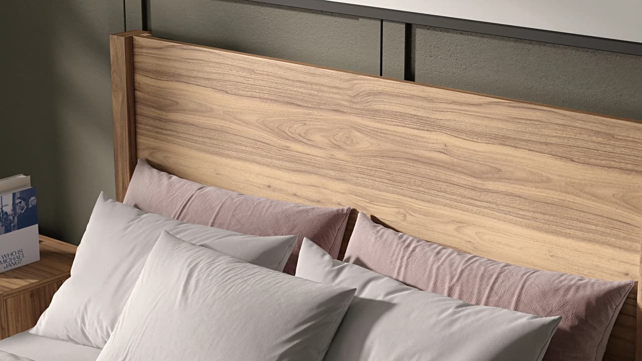 A detailed close-up of a modern 3d bed featuring a light wood headboard with a rich, natural grain pattern. The bed is styled with crisp white sleeping pillows and a long, textured dusty pink lumbar pillow, bathed in warm angled sunlight against a dark grey wall.
