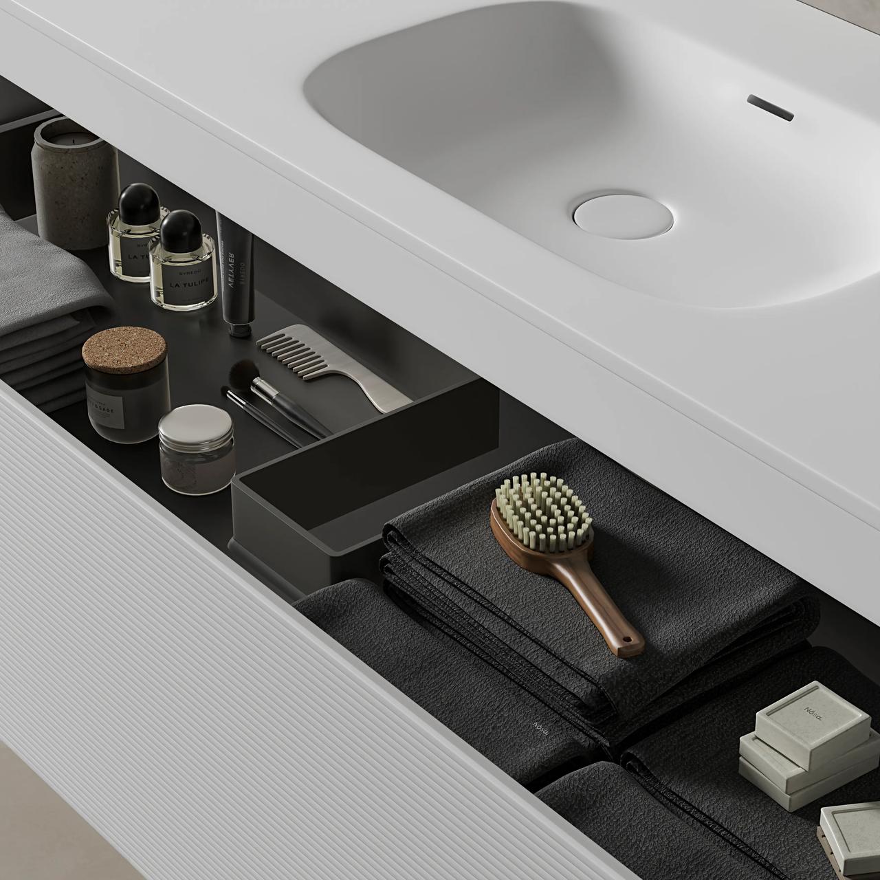 A close-up view of a white cgi bathroom vanity with an integrated sink and an open top drawer. The dark grey interior features compartments holding neatly folded dark towels, a wooden body brush, and small glass accessories.