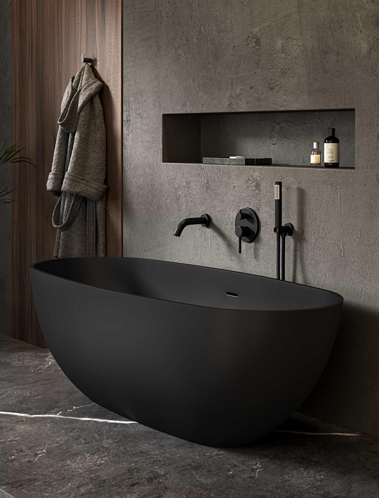 A minimalist matte black oval 3d bathtub rests on a dark marble floor against a textured grey stone wall. The moody scene features a built-in illuminated wall niche with toiletries, black wall-mounted fixtures, and a grey bathrobe hanging on a wooden panel.