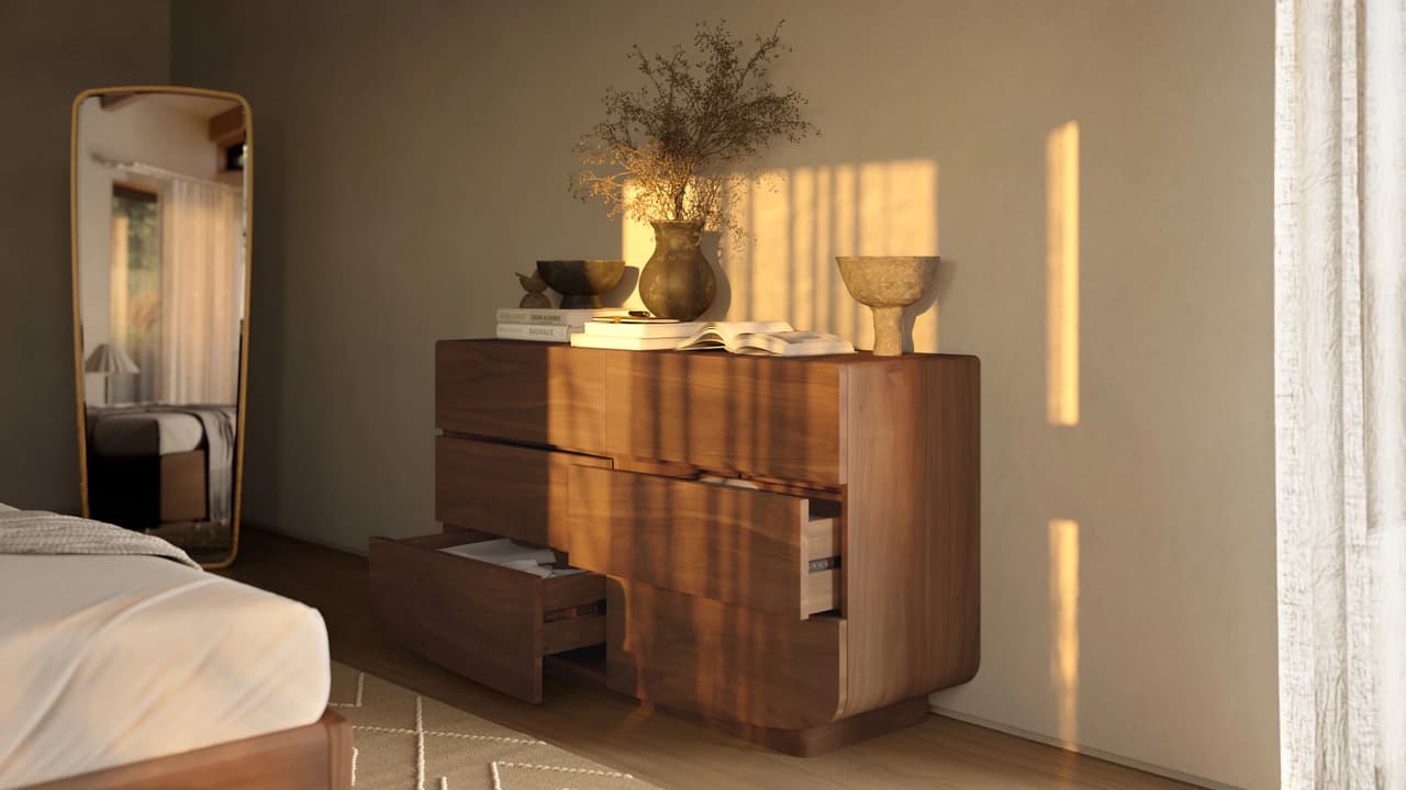 A sunlit 3d bedroom interior bathed in warm golden hour light. In the center, a modern wooden dresser with curved edges has two drawers slightly pulled open. On top of the dresser are stacked books, a rustic vase with dried branches, and a decorative ceramic bowl. To the left, a tall floor mirror reflects a window and light curtains. The edge of a bed with white linens is visible in the foreground. Strong, distinct shadows fall across the blank wall and the dresser, creating a cozy atmosphere.