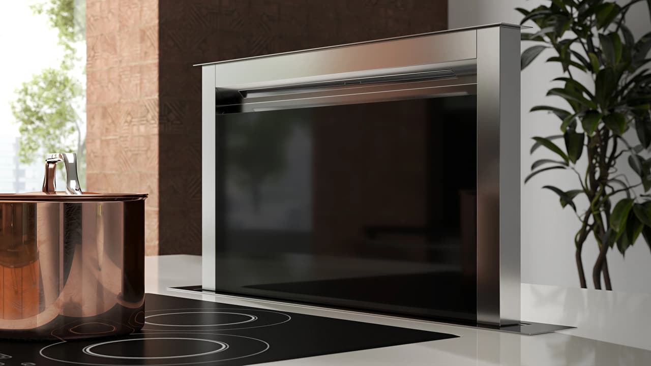 A close-up shot of a modern pop-up cgi kitchen hood featuring a stainless steel frame and a dark glass panel. It is raised from a white countertop behind a black electric cooktop with a shiny copper pot, set against a softly blurred brick wall and a potted plant.