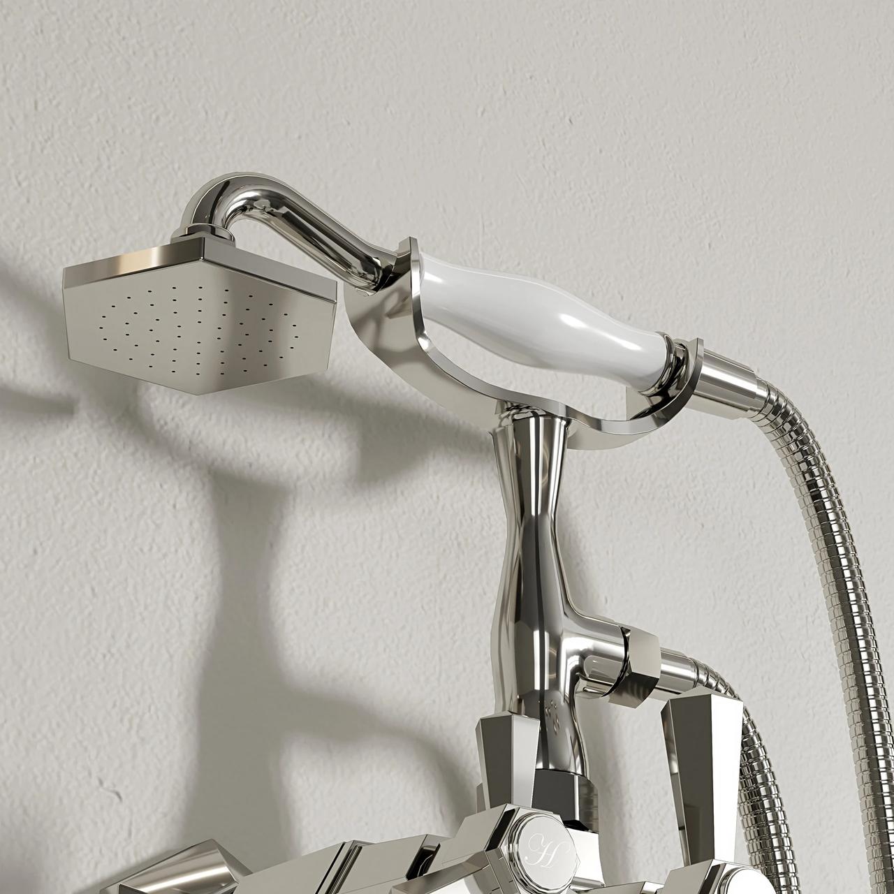 A close-up shot of a polished chrome bath faucet and handheld shower. The showerhead features a unique hexagonal face and a white ceramic handle, resting in its holder and casting a sharp shadow against a textured off-white wall.