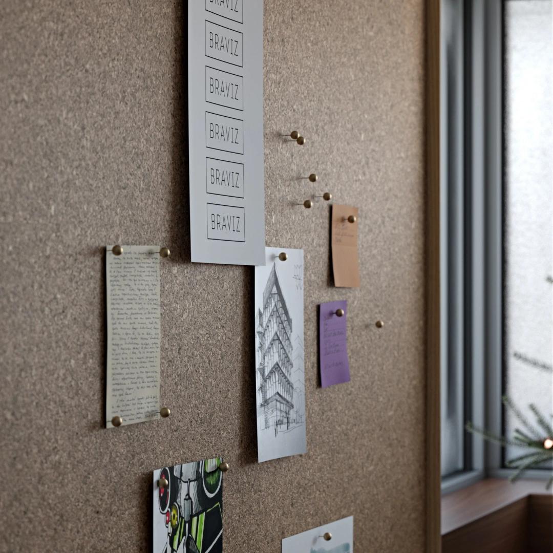 A macro shot of a textured cork 3d pinboard featuring various papers, a building sketch, and sticky notes attached with metal push pins. In the blurred background, a window frame and wooden sill add depth to the creative office scene.