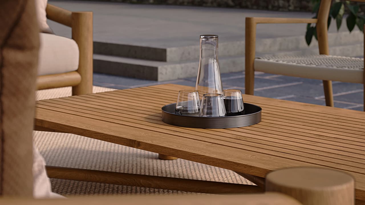 A slatted wooden outdoor 3d coffee table holds a dark round tray with a clear glass carafe and three matching cups. The blurred patio background reveals a woven rug, wooden lounge chairs, and stone steps, creating a relaxed summer atmosphere.