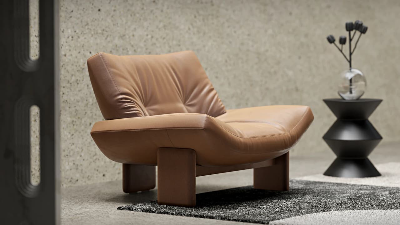 A low-profile caramel lounge 3d chair with chunky wooden legs rests on a dark rug against a lightly textured wall. Beside it, a black geometric side table holds a clear glass vase with dark flowers, creating a modern contrast.
