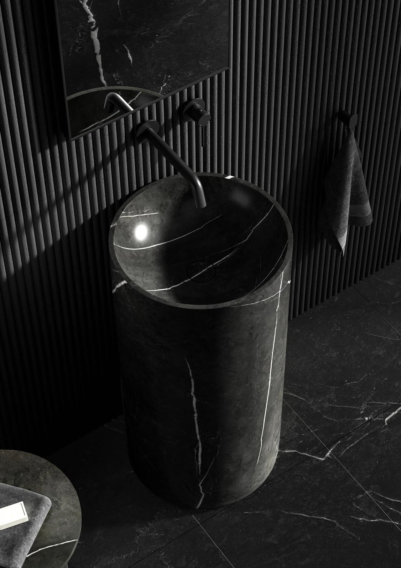 A moody bathroom interior featuring a cylindrical black marble pedestal sink with white veining. It stands on a matching dark marble floor against a black fluted wall, complete with a matte black faucet and a dark mirror reflection.