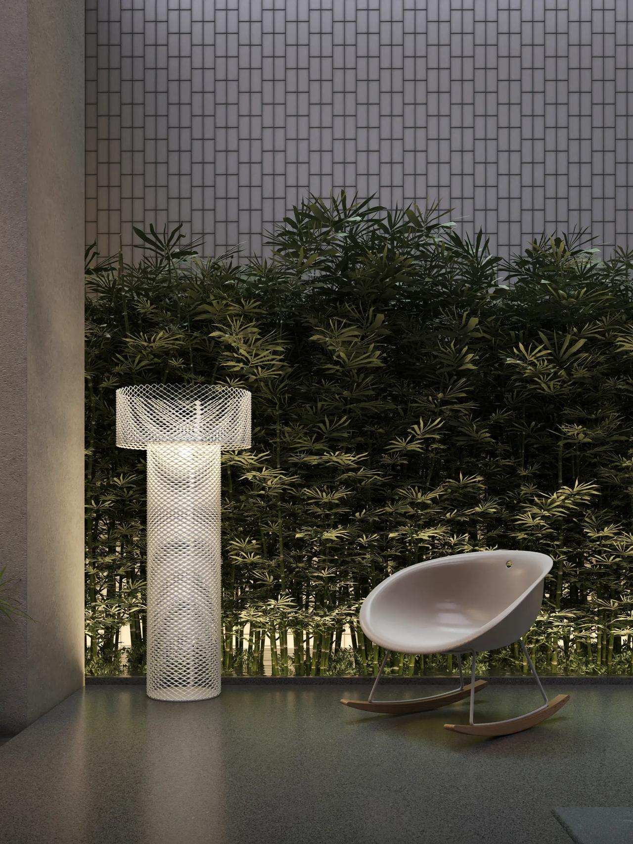 Modern rounded rocking chair set on a patio next to a tall glowing mesh floor lamp with a dense green bamboo wall in the background