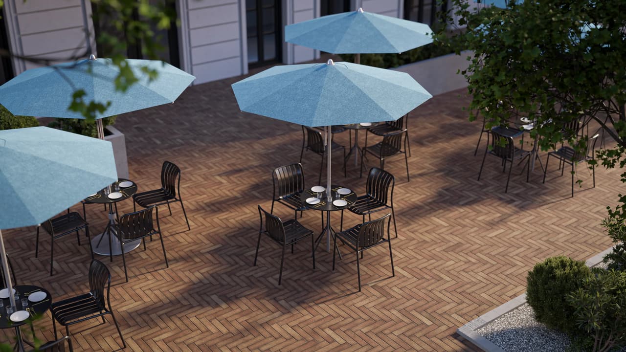 High-angle top view of an outdoor restaurant patio with paved brick flooring, round dining tables, black metal chairs, and light blue patio umbrellas