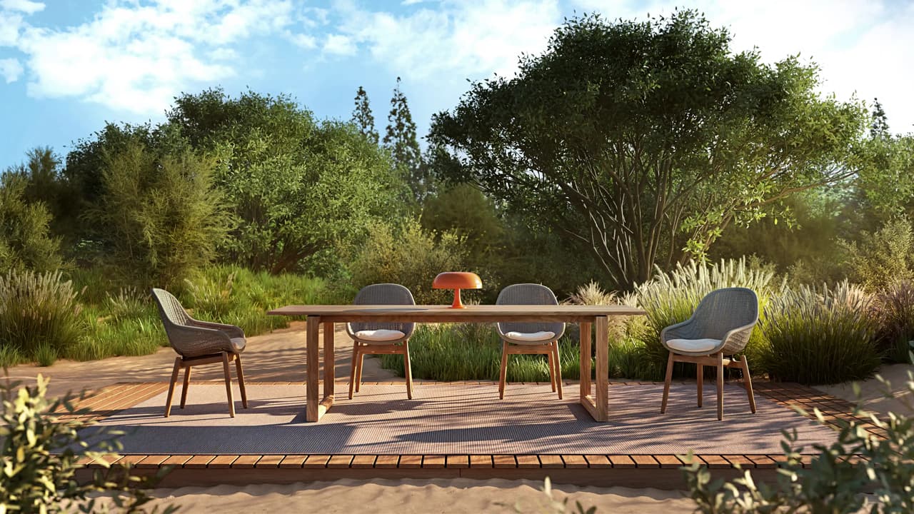 A wooden outdoor dining cgi table surrounded by four woven chairs with white cushions sits on a wooden deck. A bright orange accent lamp rests on the table, set against a beautiful background of lush green trees, tall grass, and a sunny blue sky.