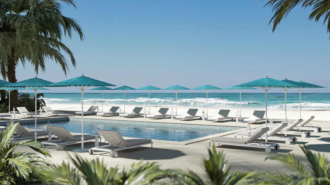 Wide view of a luxury resort pool deck with turquoise patio umbrellas, white sun loungers, and palm trees facing a tropical ocean beach